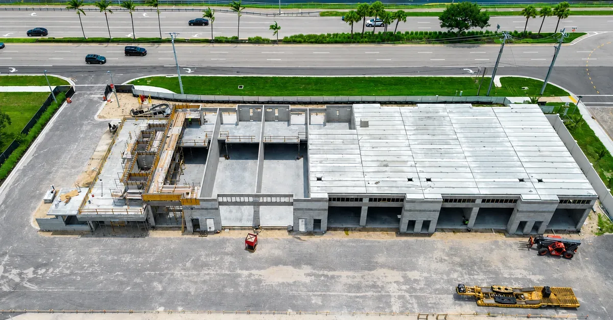 Aerial view of a commercial building under construction featuring concrete tilt-wall panels and an exposed roof deck, surrounded by heavy equipment and a bus...