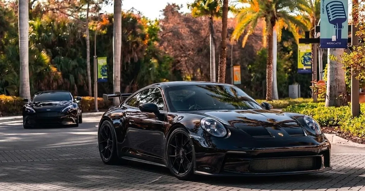 Two sleek black Porsche 911 GT3 supercars cruise through the upscale Mercato district in Naples, Florida, lined with tropical palms.